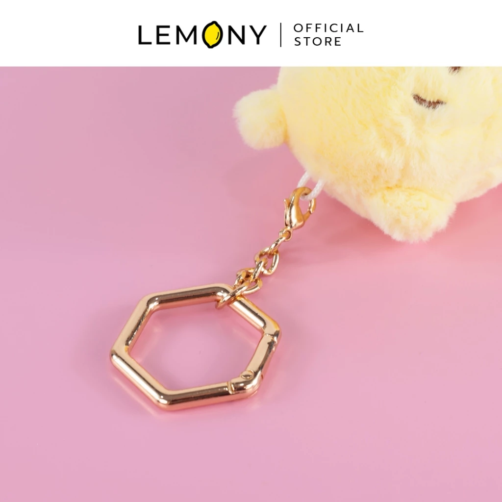 ภาพถ่ายซูมรายละเอียด LEMONY โชว์อะไหล่ห่วงกุญแจโลหะสีทองดีไซน์รูปทรงหกเหลี่ยมพรีเมียม เชื่อมต่อด้วยสายโซ่สั้นและตัวล็อกก้ามปูเข้ากับสายผ้าสีขาวบนหัวตุ๊กตาหมีพูห์ขนนุ่ม บนพื้นหลังสีชมพูพาสเทล