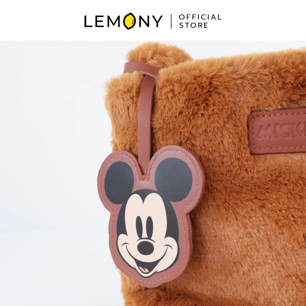ภาพถ่ายซูมใกล้โชว์จี้ห้อยหนังสีน้ำตาลตัดขอบรูปหัวมิกกี้เมาส์ (Mickey Mouse) หน้าตรงยิ้มแฉ่ง ประดับด้วยสายหนังคล้องติดกับขอบกระเป๋าขนนุ่มสีน้ำตาล ข้างกันมีป้ายหนังปั๊มอักษรคำว่า MICKEY