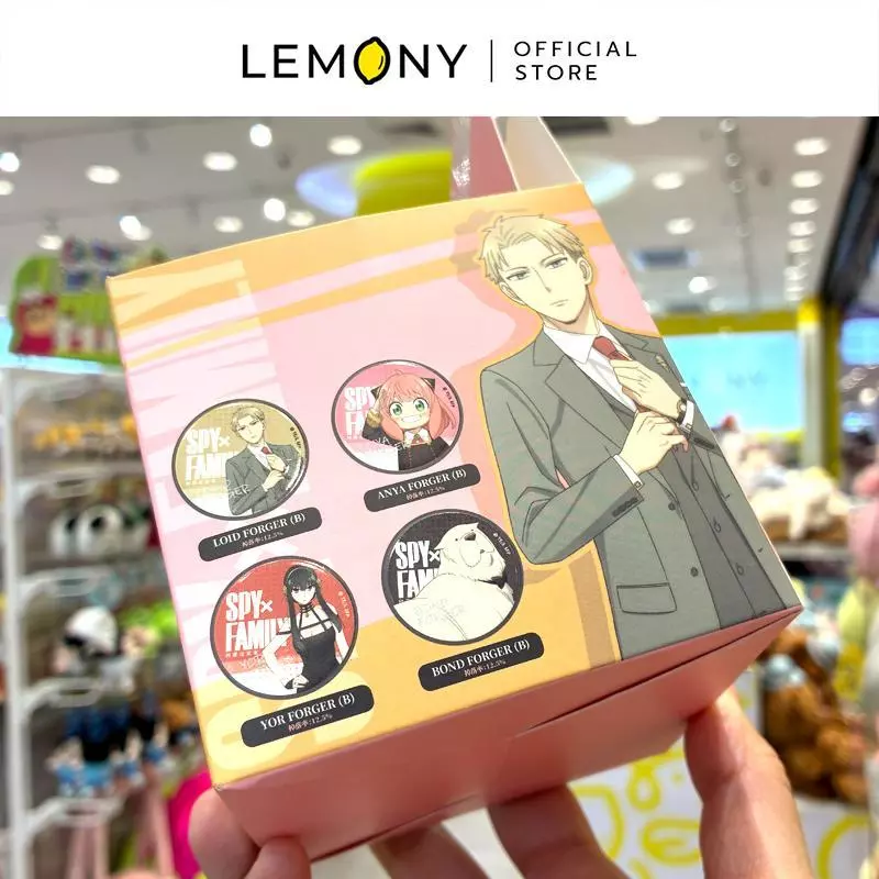 โทนสีโอรสและชมพูพาสเทล พิมพ์ลายกราฟิกตัวละครลอยด์ ฟอร์เจอร์ (Loid Forger) ในชุดสูทสากลสีเทาสุดเนี้ยบ ข้าง ๆ แสดงรูปภาพตัวอย่างเข็มกลัดทรงกลมดีไซน์เวอร์ชัน B จำนวน 4 ลาย ได้แก่ ลอยด์, อาเนีย, ยอร์ และสุนัขบอนด์ ฉากหลังเป็นชั้นวางของเล่นเบลอในร้าน ด้านบนสุดระบุโลโก้แบรนด์ LEMONY Official Stor