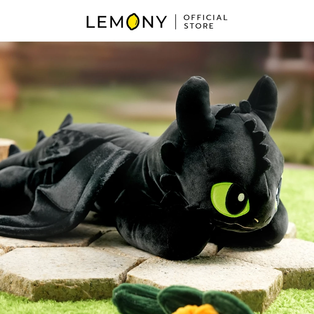 ภาพถ่ายระยะใกล้จากมุมเฉียงด้านขวา เผยใบหน้าตุ๊กตาผ้าเขี้ยวกุด (Toothless) มังกรเพลิงนิลสีดำสนิทหนานุ่ม โชว์งานปักดวงตาสีเขียวสดใสโตเด่นชัด กำลังนอนหมอบราบอยู่บนแผ่นหินปูพื้นทรงหกเหลี่ยมท่ามกลางสนามหญ้าสีเขียว ด้านบนสุดพิมพ์แถบป้ายตราแบรนด์ LEMONY Official Store