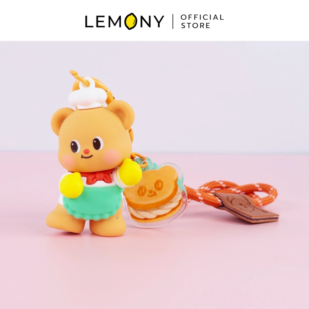 ภาพถ่ายสไตล์ไลฟ์สไตล์ จัดวางพวงกุญแจโมเดลหมีเนย Butterbear สวมหมวกเชฟสีขาวและผ้ากันเปื้อนสีฟ้ามิ้นต์ ยืนโพสต์ท่าน่ารักคู่กับจี้อะคริลิกลายแซนด์วิชเนย พ่วงสายคล้องข้อมือเชือกถักสีส้มอิฐ วางบนพื้นหลังสีชมพูละมุน ด้านบนสุดระบุโลโก้แบรนด์ LEMONY Official Store
