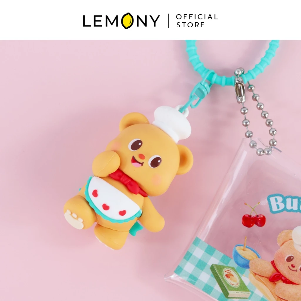 ภาพถ่ายสไตล์ไลฟ์สไตล์ โฟกัสชิ้นงานโมเดลยางซิลิโคนน้องหมีเนย Butterbear หน้าตรงอมยิ้ม สวมหมวกเชฟและผ้ากันเปื้อนขอบฟ้ามิ้นต์ลายหัวใจ พ่วงซองกระเป๋าใสทำขนม และสายห้อยซิลิโคนสีฟ้ามิ้นต์ ฉากหลังสีชมพูหวานละมุน ด้านบนสุดระบุโลโก้แบรนด์ LEMONY Official Store
