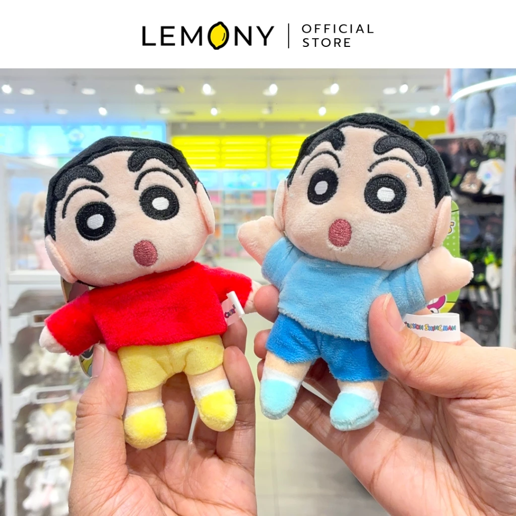 ภาพถ่ายสไตล์ไลฟ์สไตล์ เผยมุมมองสองมือที่กำลังถือโชว์พวงกุญแจตุ๊กตาผ้าชินจังหน้าตรง 2 ลาย ฝั่งซ้ายคือดีไซน์ชุดคลาสสิกเสื้อสีแดงกางเกงสีเหลือง ฝั่งขวาคือดีไซน์เสื้อแขนสั้นและกางเกงขาสั้นสีฟ้าสดใส ตัวตุ๊กตาทำจากผ้าขนสั้นเนื้อนุ่มเย็บปักรายละเอียดประณีต ด้านบนสุดระบุโลโก้แบรนด์ LEMONY Official Store