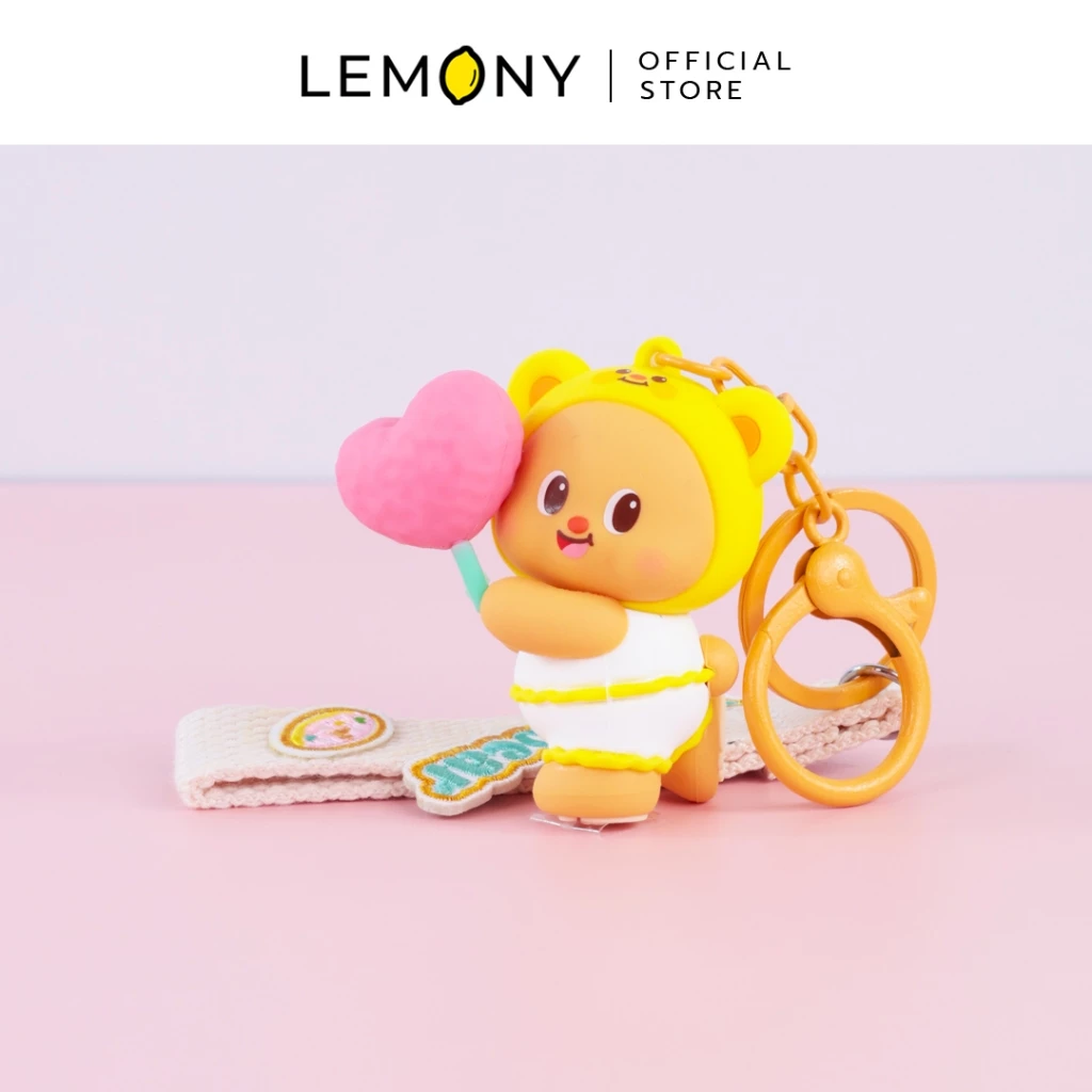 ภาพถ่ายสไตล์ไลฟ์สไตล์ จัดวางพวงกุญแจโมเดลน้องหมีเนย Butterbear สวมหมวกฮู้ดหมีสีเหลืองถือสายไหมรูปหัวใจสีชมพู วางคู่กับสายคล้องผ้าแคนวาสและห่วงตะขอเกี่ยวสีเหลืองสดใส บนพื้นหลังสีชมพูละมุน ด้านบนสุดระบุโลโก้แบรนด์ LEMONY Official Store