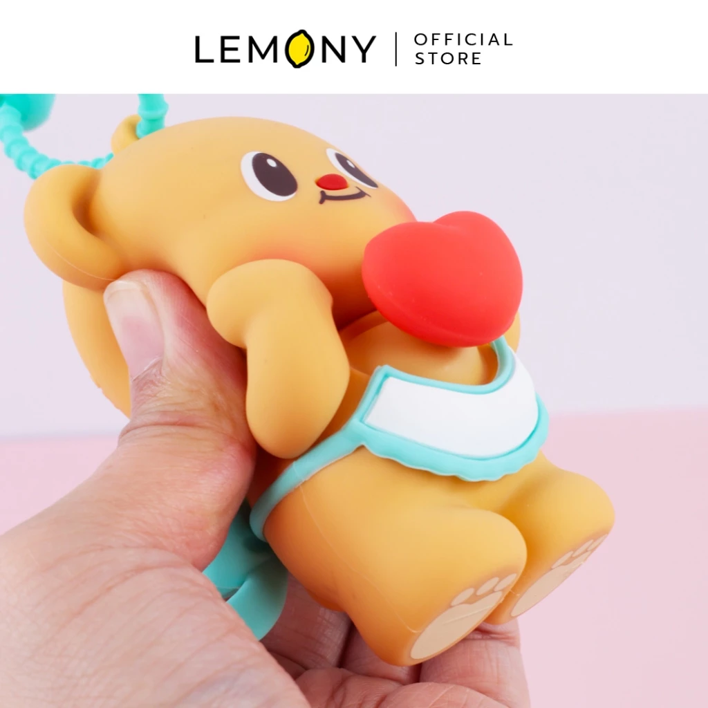 ภาพถ่ายระยะใกล้ เผยมุมมองมือซ้ายกำลังจับบีบพวงกุญแจโมเดลน้องหมีเนย Butterbear สีเหลืองนวล เผยให้เห็นความนุ่มนิ่มยืดหยุ่นของวัสดุยางซิลิโคนคุณภาพดี สวมผ้ากันเปื้อนสีขาวขอบฟ้ามิ้นต์ กอดดวงใจสีแดงสดใส ฉากหลังสีชมพูละมุน ด้านบนสุดระบุโลโก้แบรนด์ LEMONY Official Store