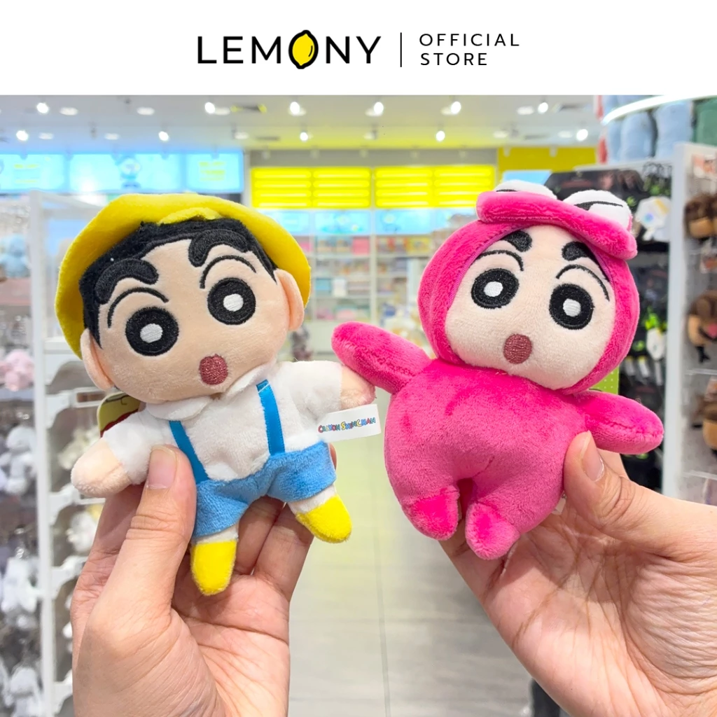 ภาพถ่ายสไตล์ไลฟ์สไตล์ เผยมุมมองสองมือที่กำลังถือโชว์พวงกุญแจตุ๊กตาผ้าชินจังตัวนุ่มนิ่มพร้อมกัน 2 แบบ ฝั่งซ้ายคือชินจังสวมหมวกสีเหลืองเสื้อขาวเอี๊ยมสีฟ้า ฝั่งขวาคือชินจังในชุดมาสคอตไดโนเสาร์วานิยามาซังสีชมพูเข้มทำหน้าตาตื่นเต้น ฉากหลังเป็นบรรยากาศชั้นวางสินค้าภายในร้านค้า ด้านบนสุดระบุโลโก้แบรนด์ LEMONY Official Store