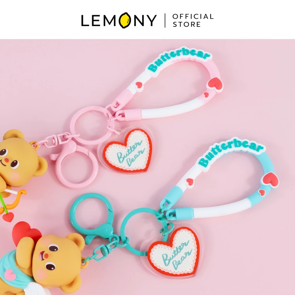 ภาพถ่ายระยะใกล้ ซูมรายละเอียดสายคล้องข้อมือยางซิลิโคนปั๊มนูนข้อความ Butterbear แต่งรูปดวงใจสีแดงสองเฉดสี (สีชมพูพาสเทลและสีฟ้ามิ้นต์) พ่วงจี้อะคริลิกใสทรงหัวใจลายขอบลูกไม้สกรีนข้อความ Butter Bear ฉากหลังสีชมพูละมุน ด้านบนสุดระบุโลโก้แบรนด์ LEMONY Official Store