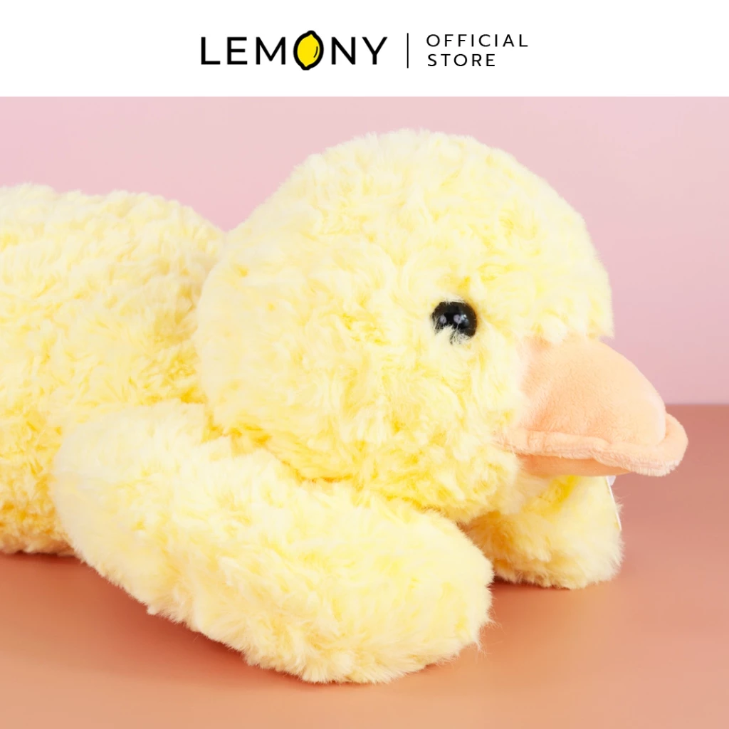 ภาพถ่ายระยะใกล้โชว์รายละเอียดความนุ่มฟูของตุ๊กตาเป็ดสีเหลืองสดใส เผยผิวสัมผัสผ้าขนสัตว์สังเคราะห์เส้นยาวเป็นปุ่มนุ่มมือ แต่งดวงตาสีดำกลมโตและปากแบนสีส้มพาสเทลทำจากผ้าขนนุ่มสั้น วางบนพื้นผิวสีส้มฉากหลังสีชมพูละมุน ด้านบนสุดระบุโลโก้แบรนด์ LEMONY Official Store